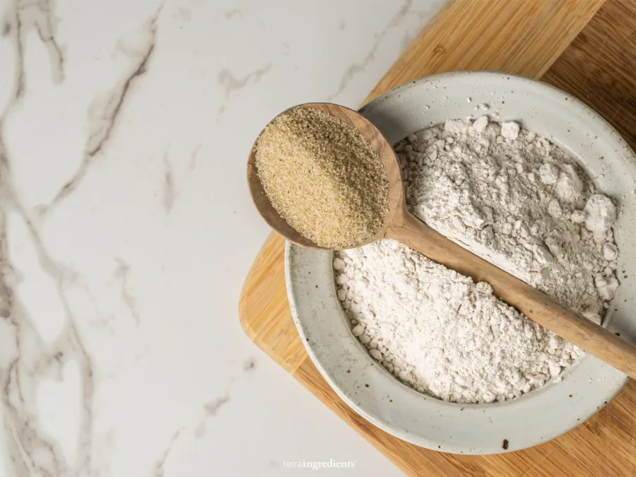 A wooden spoon with raw fonio grain sits upon a bowl of fonio flour.