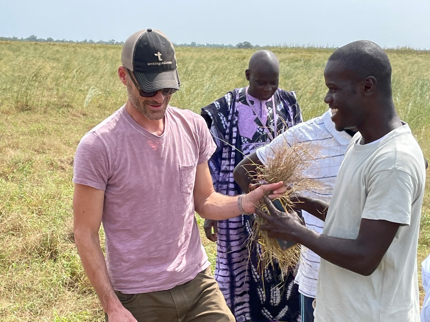 Two men smile while handing over a bushel of harvested fonio