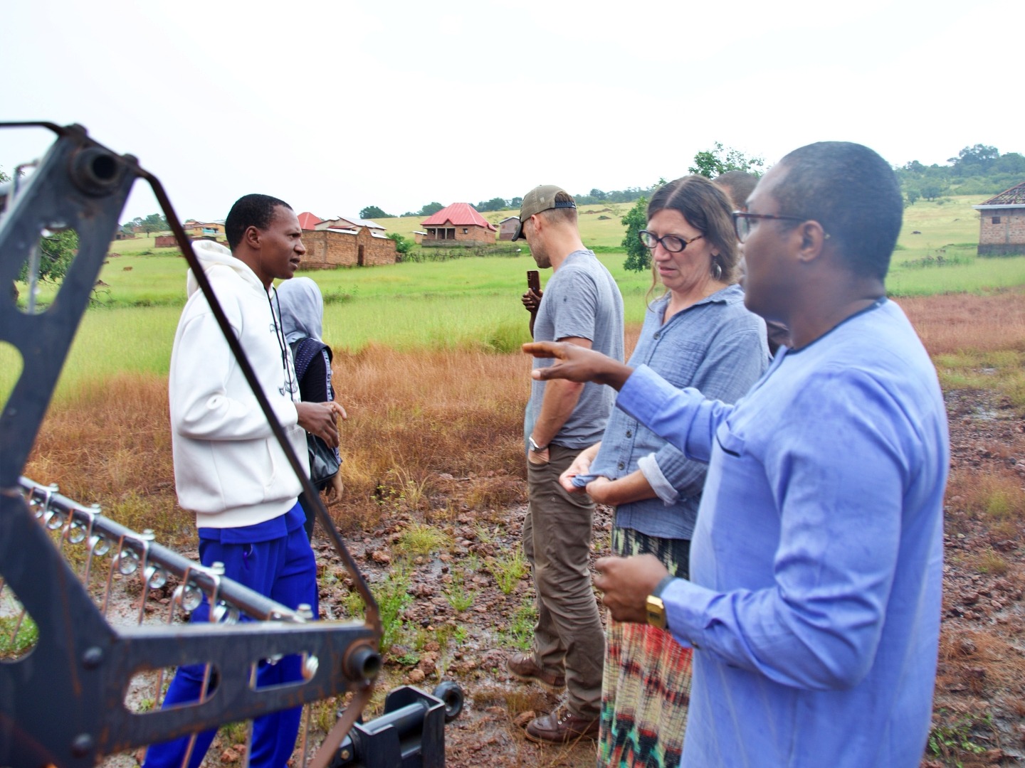 A man in blue traditional African clothing speaks with a woman next to farm equipment.