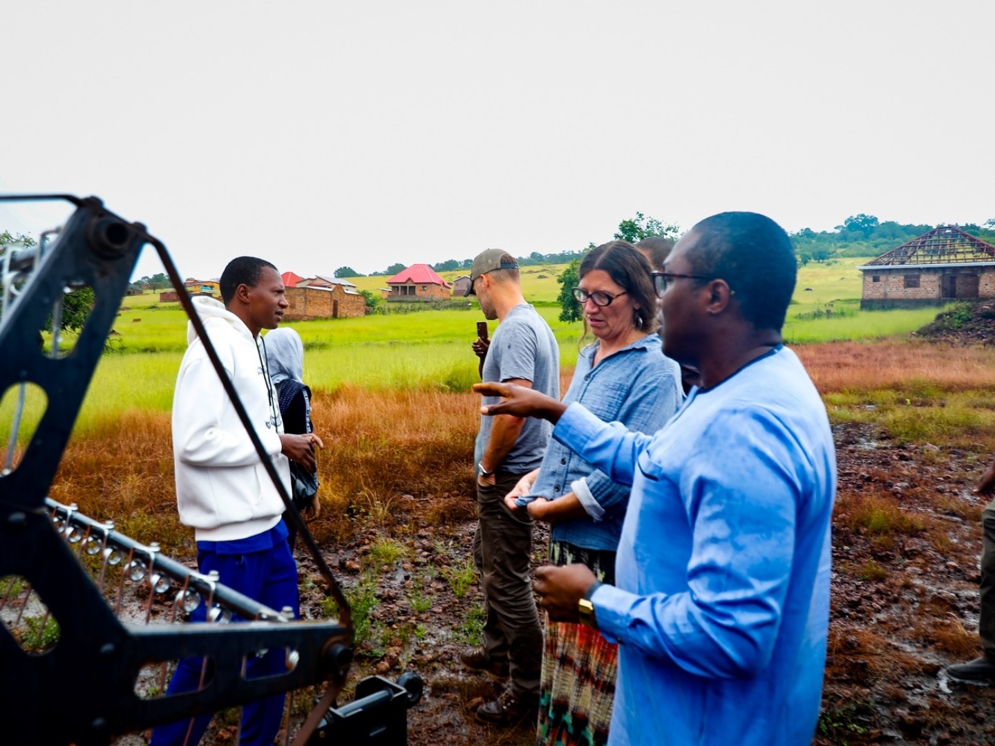 A man in blue traditional African clothing speaks with a woman next to farm equipment.