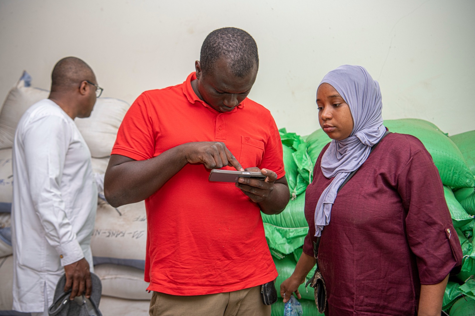 A man and a woman are looking at a phone while another man inspects bags of grain in the background